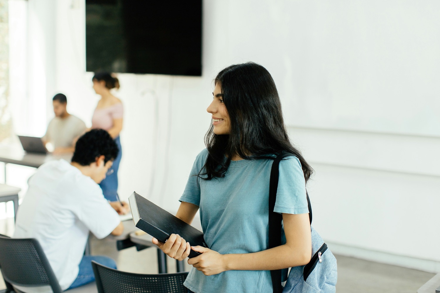 A team member with a notebook and backpack in a bright office, colleagues working behind her.